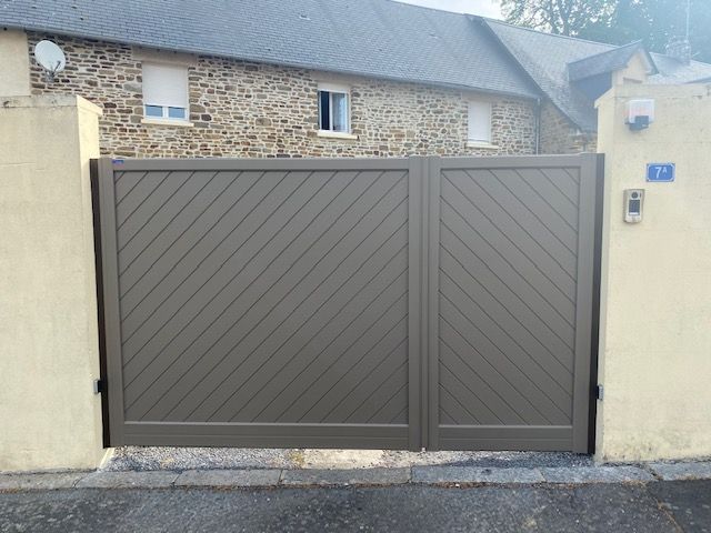 Portail à lattes diagonales marron entre des piliers beiges, devant une maison en pierre.