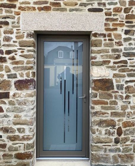 Porte grise avec lignes gravées, encastrée dans un mur en pierre, avec linteau en granit clair.