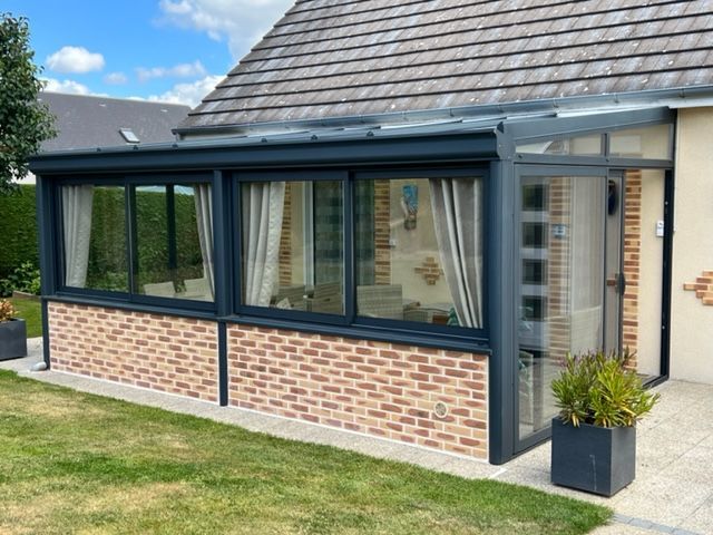 Ajout d'une véranda avec base en brique, fenêtres en verre et charpente gris foncé, attachée à une maison.