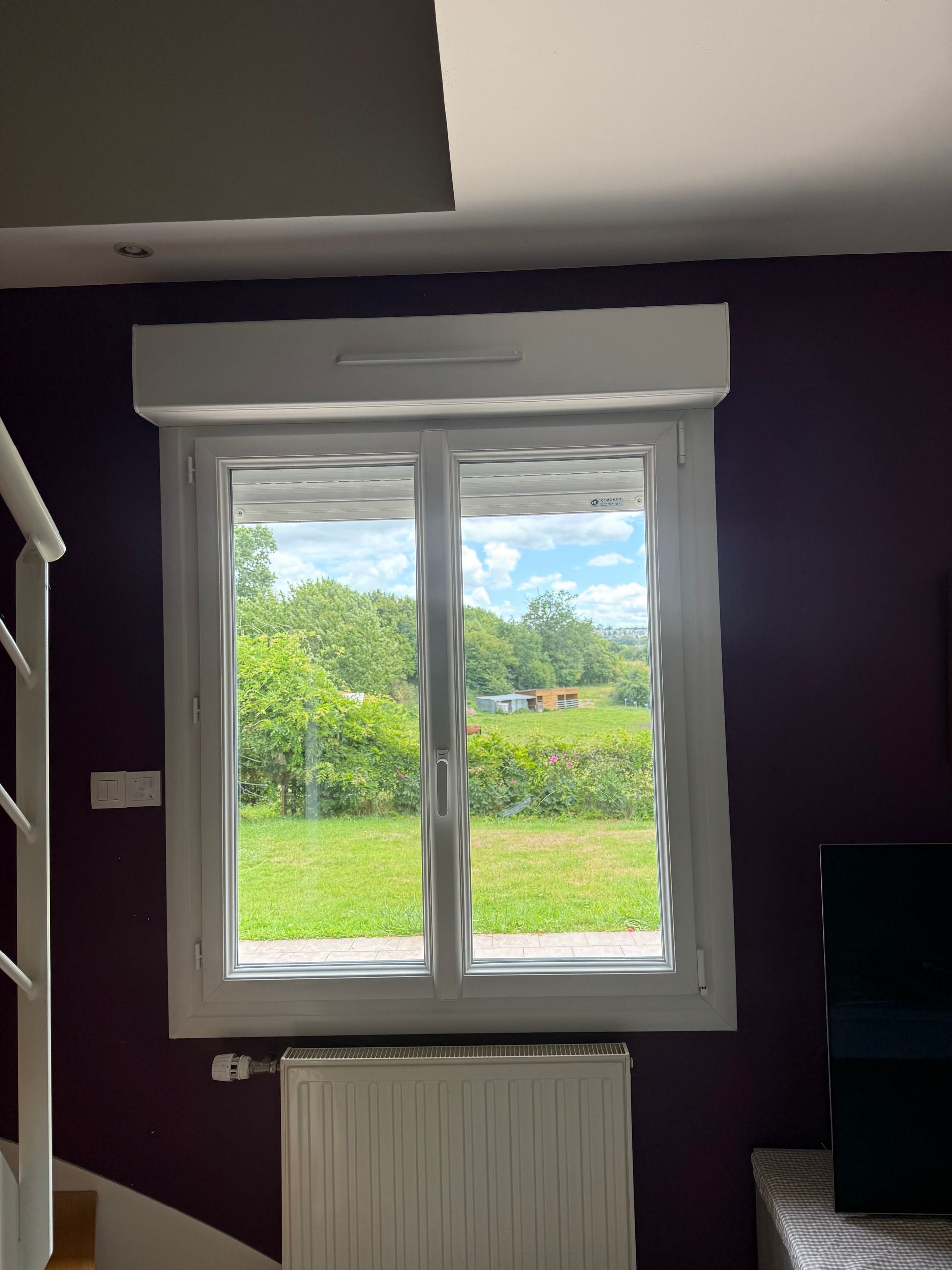 Fenêtre avec vue sur un champ verdoyant et des arbres. Cadre blanc, mur violet, radiateur blanc et store partiellement fermé.