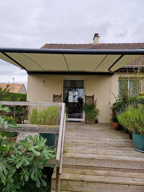 Terrasse en bois plantée de plantes, menant à une maison avec un auvent au-dessus de la porte. Ciel nuageux.