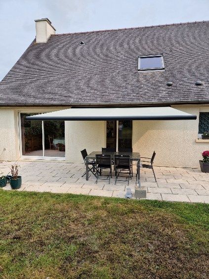 Terrasse arrière avec auvent rétractable, table et chaises. Une maison au toit sombre en arrière-plan.