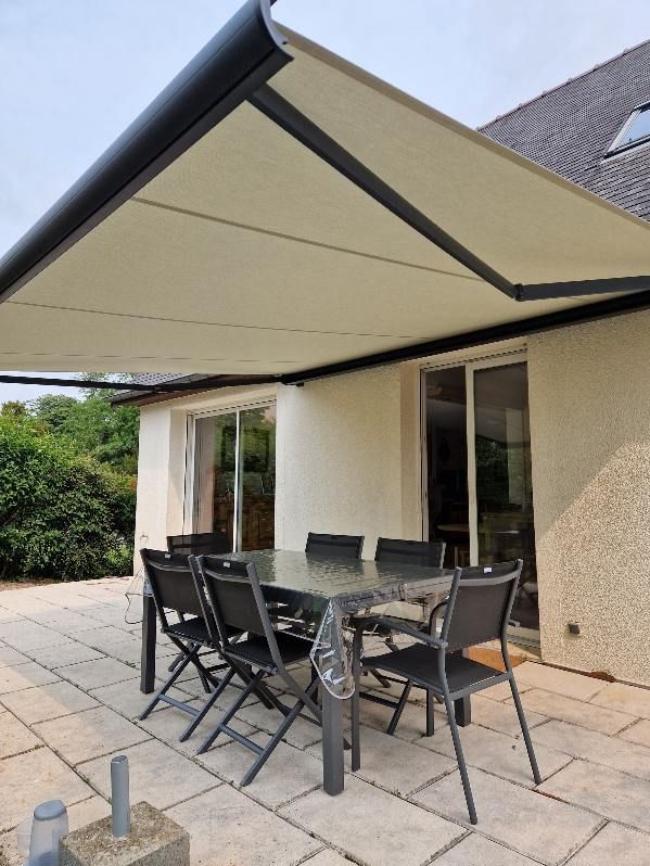 Patio avec table à manger, chaises sous un auvent rétractable beige, à côté d'une maison avec portes coulissantes en verre.