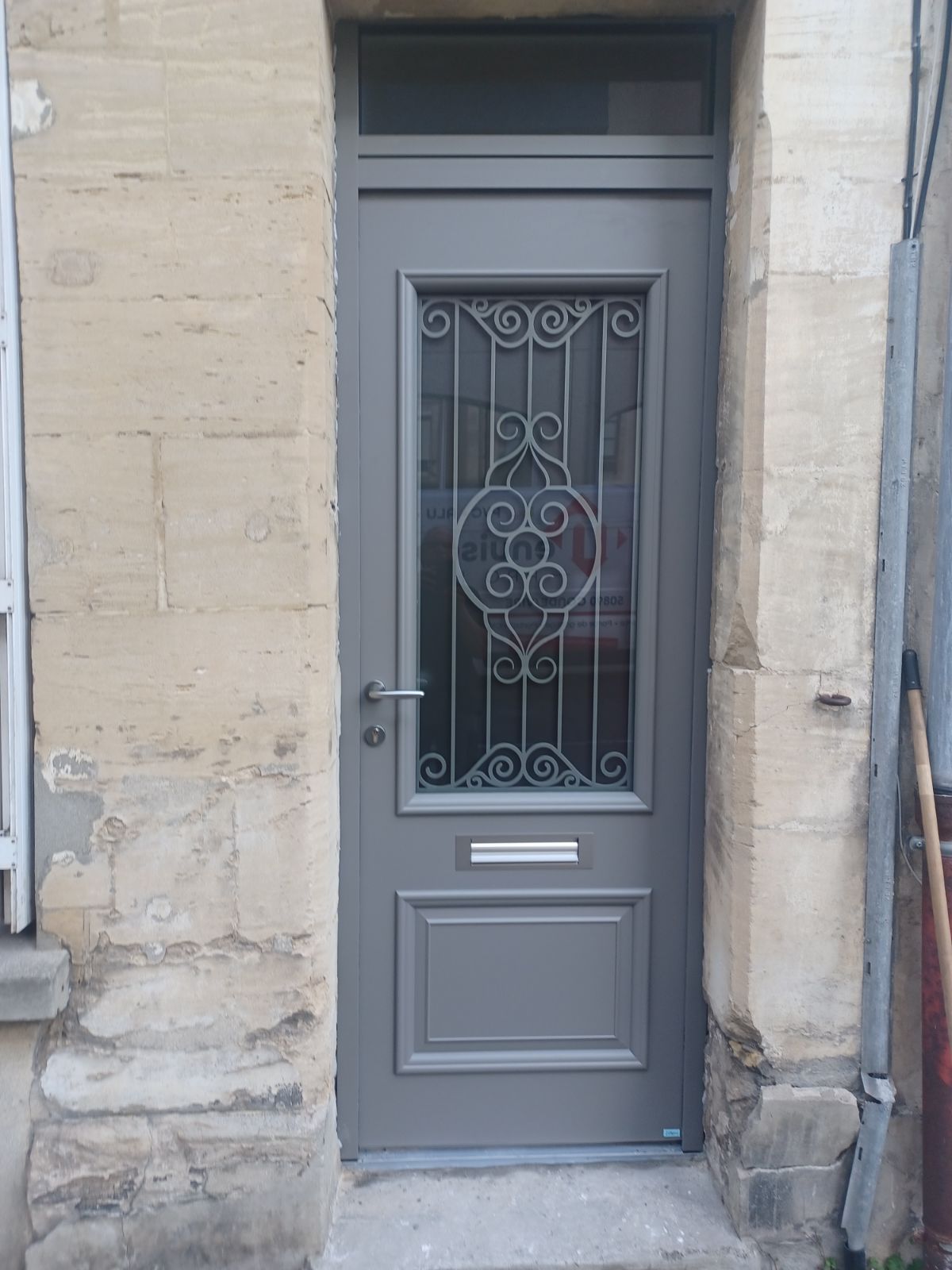 Porte grise avec panneau de verre orné, située dans un bâtiment en pierre.