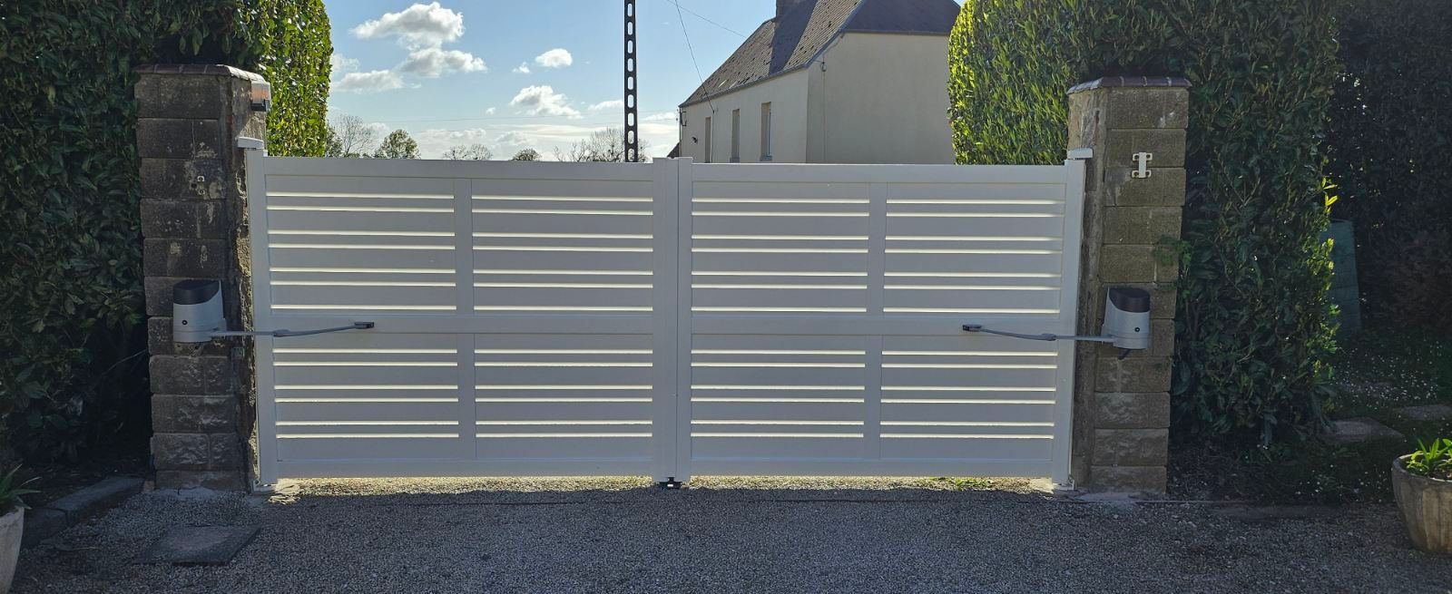 Portail automatique blanc, lames horizontales, piliers en briques, allée en gravier, feuillage vert.