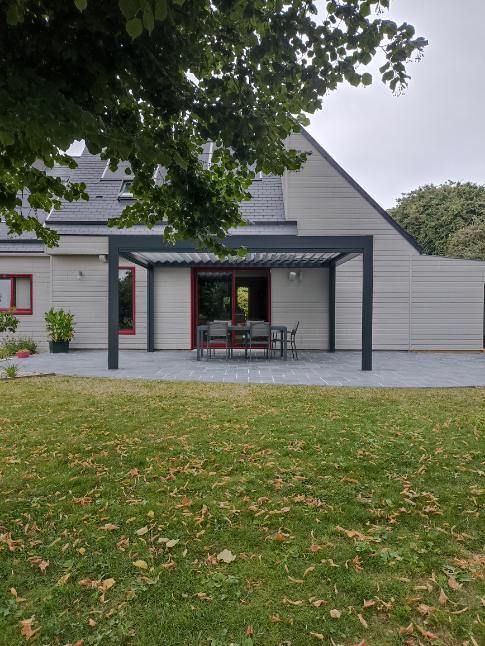 Une pergola grise sur un patio avec mobilier d'extérieur ; maison en arrière-plan, herbe verte au premier plan.