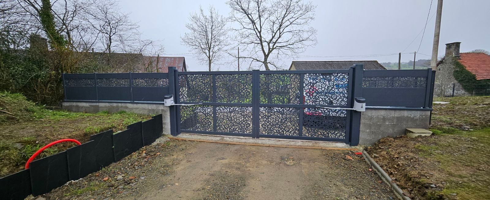 Un portail décoratif en métal noir avec une clôture assortie sur une allée en gravier. Arbres nus et ciel nuageux en arrière-plan.