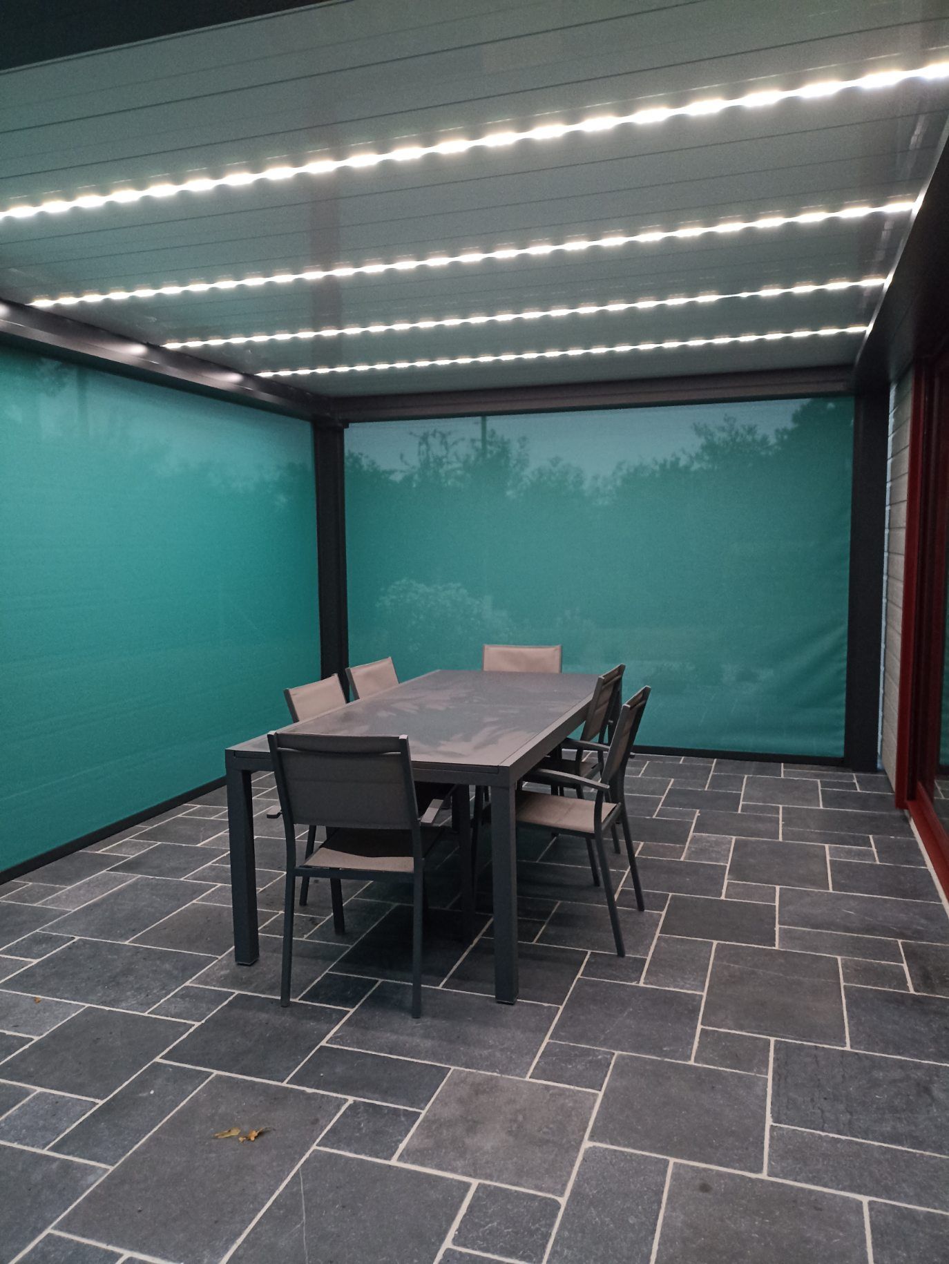 Terrasse avec table et chaises sous un auvent orné de rideaux bleu sarcelle. Luminaires au plafond.