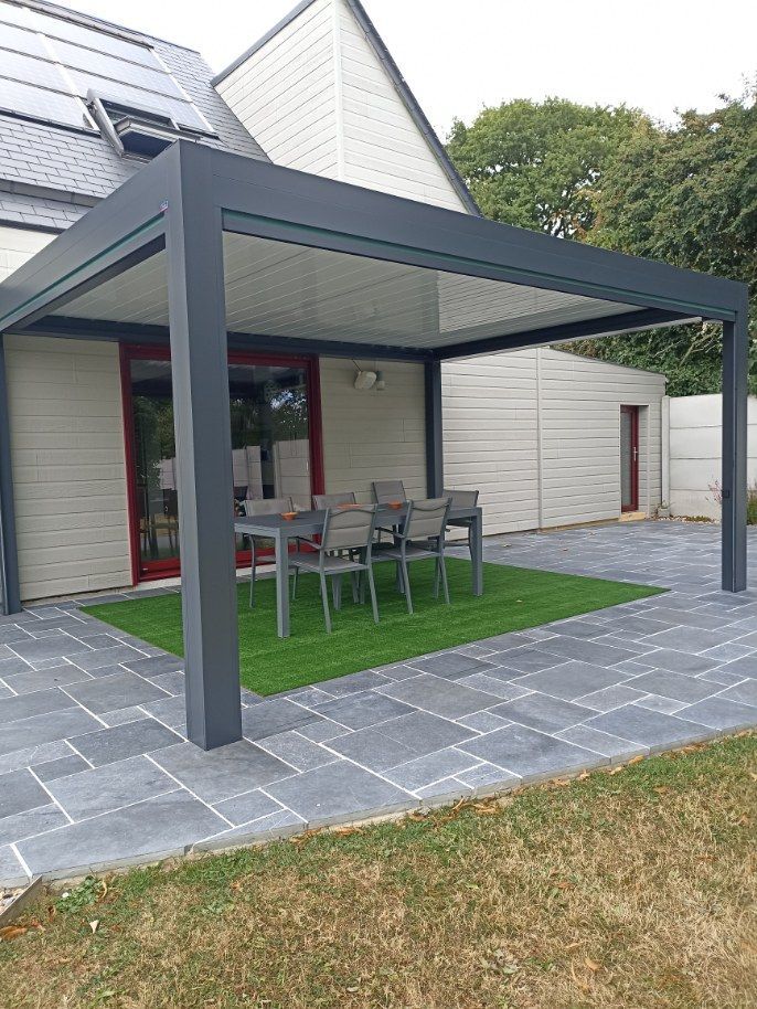 Pergola grise sur mobilier de patio sur gazon artificiel, adjacente à une maison.