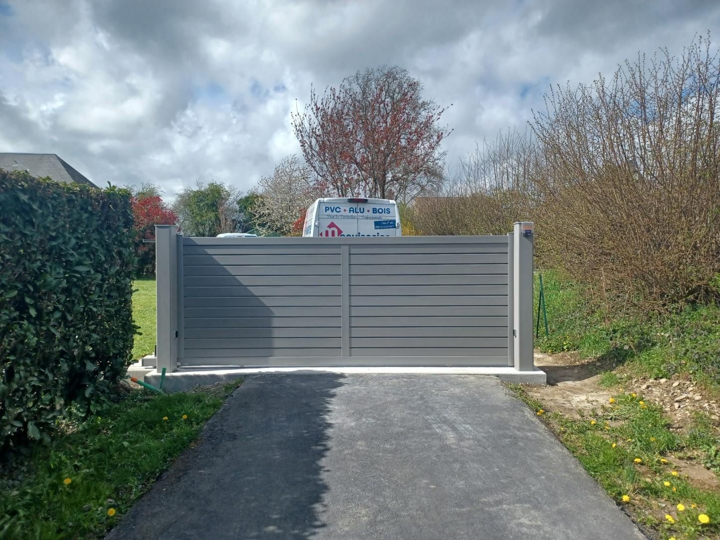 Portail à lattes horizontales gris sur une allée, avec un véhicule visible derrière.