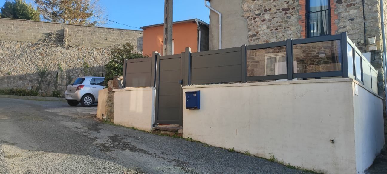 Une voiture argentée garée près d'un chemin de gravier, des murs blancs et une clôture grise avec une boîte aux lettres bleue.