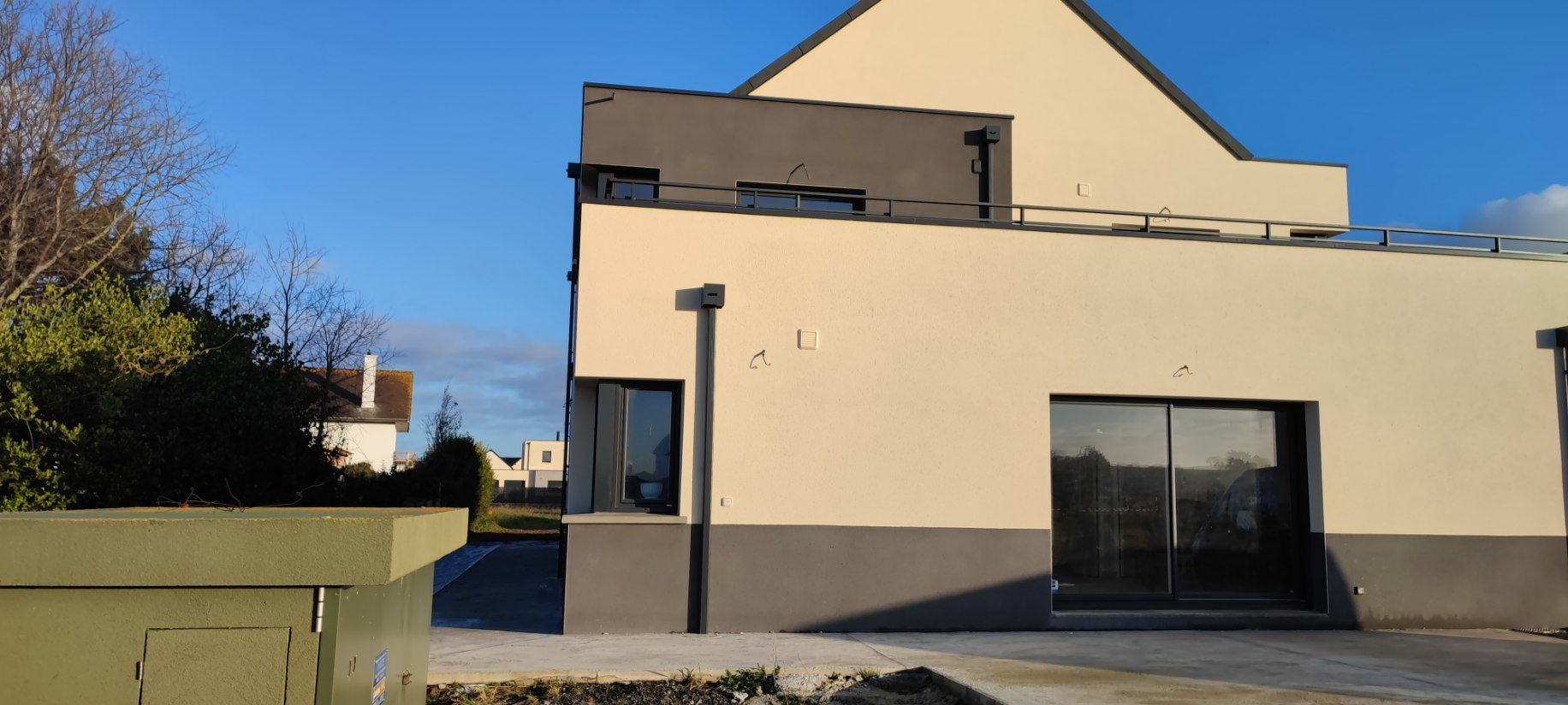 Extérieur de maison moderne, murs crème, accents gris foncé, grandes portes coulissantes en verre, ciel bleu.