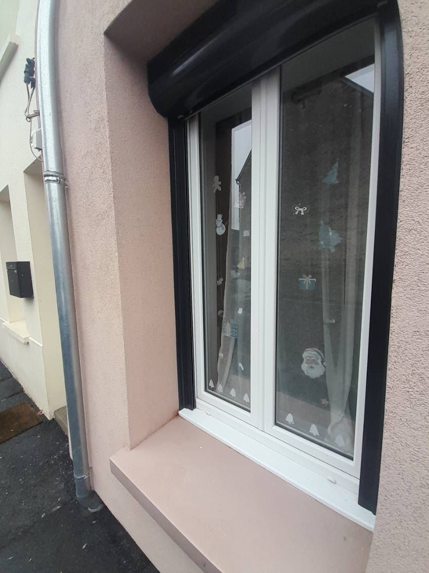 Fenêtre avec un volet noir, un cadre blanc et un mur extérieur rose.