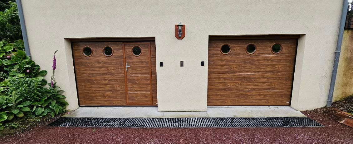 Deux portes de garage en bois avec des fenêtres rondes, une crête au-dessus et une cour en gravier rouge.