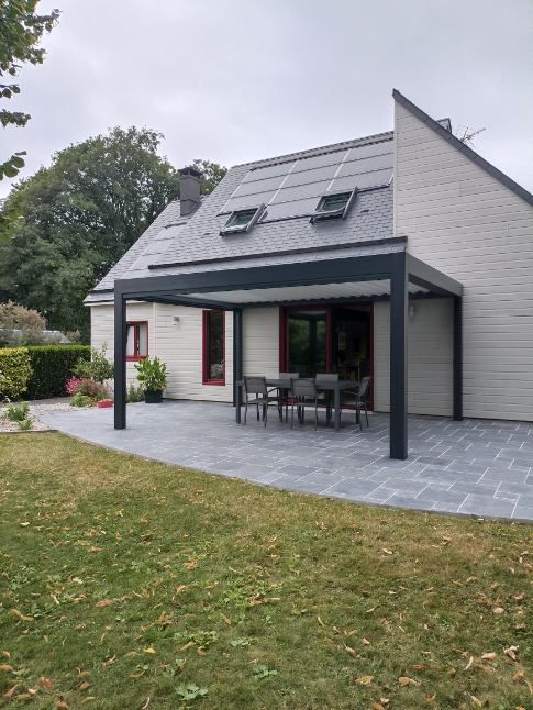 Patio arrière avec pergola foncée, table et chaises, à côté d'une maison de couleur claire avec panneaux solaires.