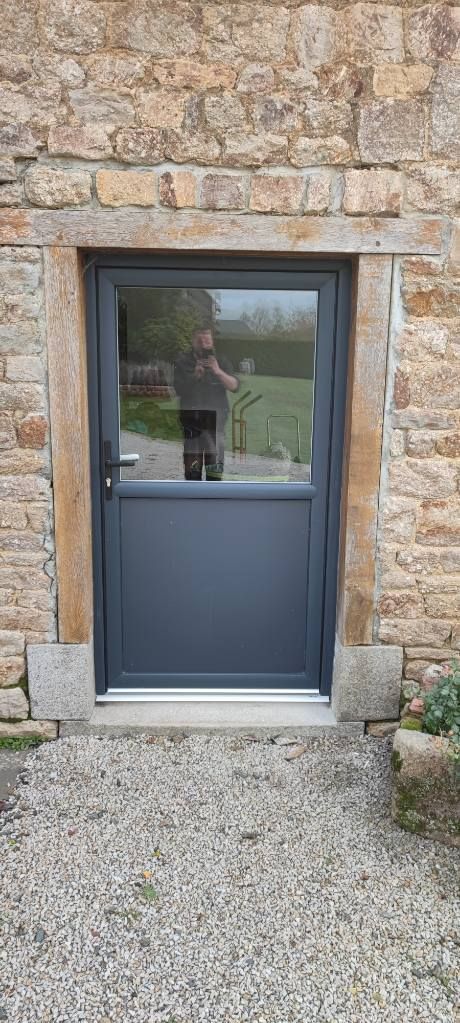 Une porte gris foncé encastrée dans un mur de pierre. Un chemin de gravier y mène.