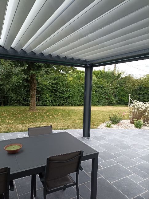 Patio avec table et chaises noires, sous un toit rétractable, donnant sur une cour verdoyante.