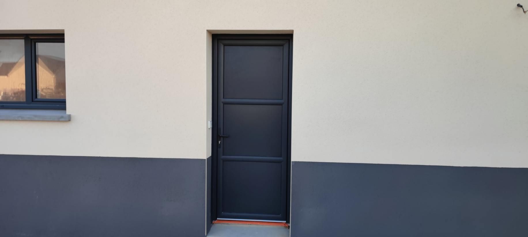Une porte en métal noir centrée sur un mur blanc et gris avec une fenêtre à gauche.