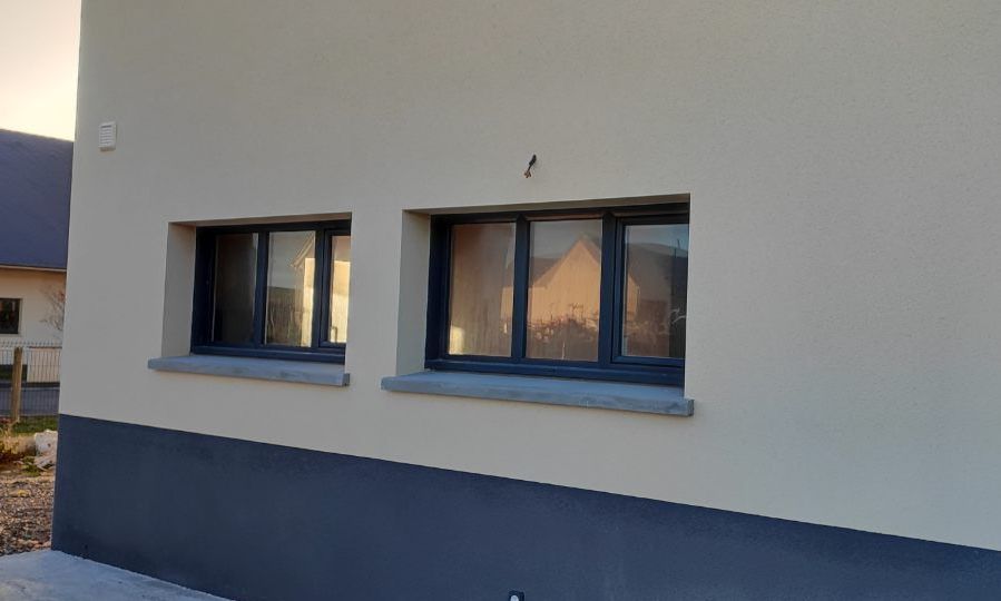 Deux fenêtres à cadre gris sur une maison gris clair avec des fondations gris foncé.