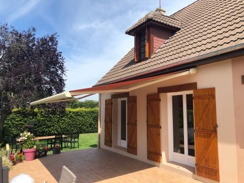 Maison beige avec auvent de toit rouge, volets en bois et pelouse verte par une journée ensoleillée.