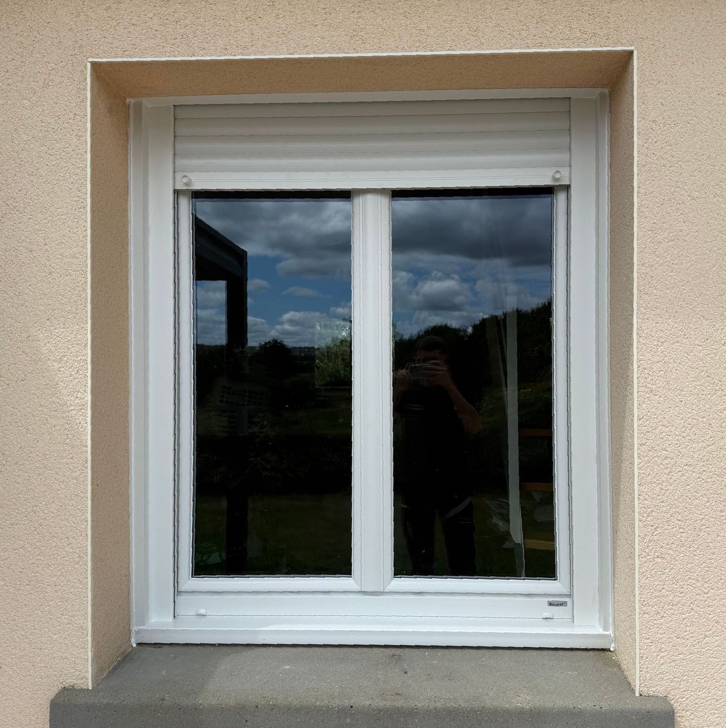 Fenêtre avec cadre blanc et store fermé, reflétant un ciel nuageux et une personne tenant un téléphone.