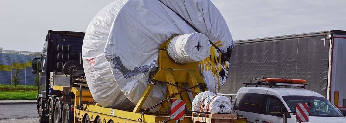Un gran objeto cilíndrico envuelto en una plataforma de camión es seguido por un coche de policía en una carretera.