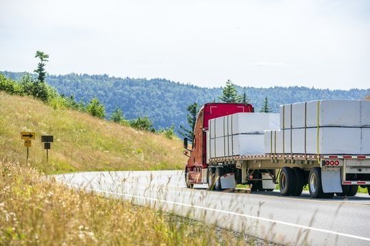Camión semirremolque rojo que transporta carga blanca apilada en un camino sinuoso con una colina cubierta de hierba y un bosque al fondo.