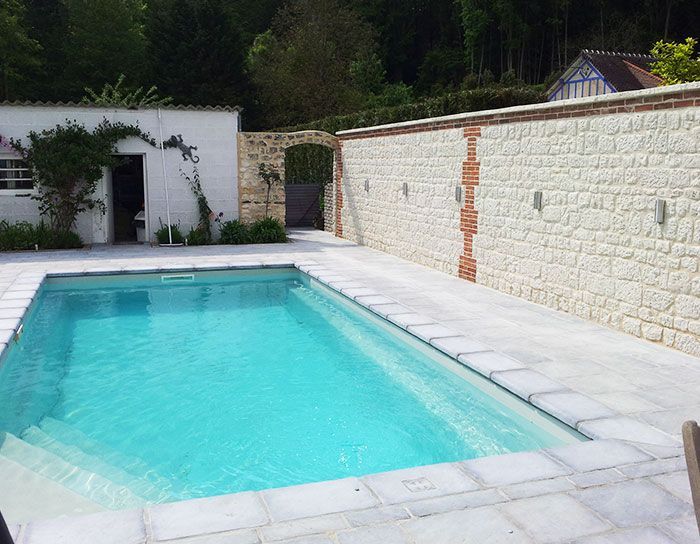Piscine dans le jardin, bordée d'un muret en pierre et d'une terrasse blanche, avec une eau turquoise et des arbres en arrière-plan.