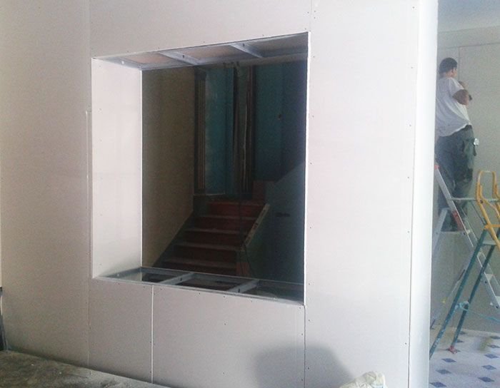 Mur blanc avec une fenêtre carrée donnant sur une cage d'escalier sombre ; un ouvrier sur une échelle, au bord droit.