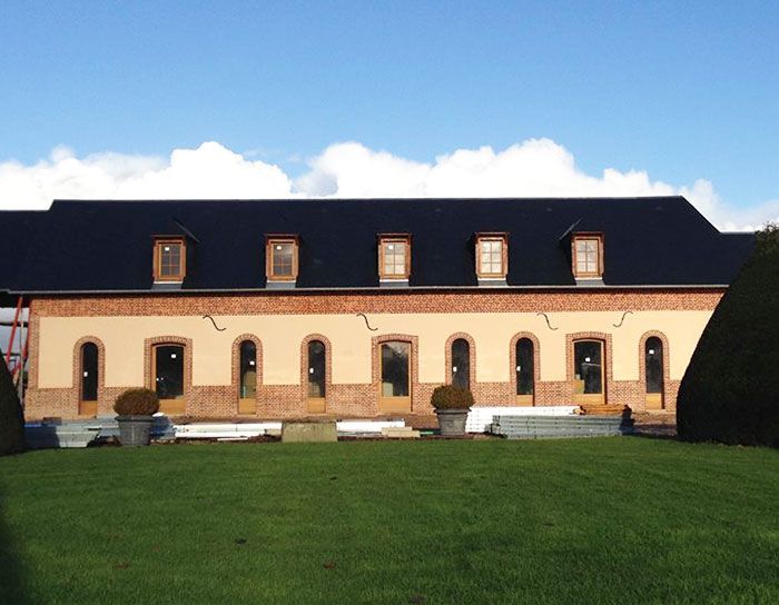 Longue maison beige au toit noir et aux lucarnes, se détachant sur une pelouse verte sous un ciel bleu.