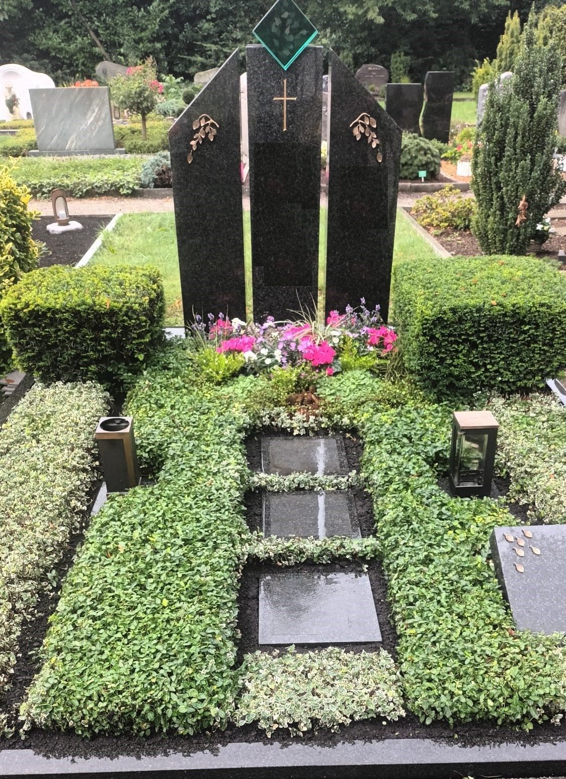 Grab mit schwarzem Grabstein, umgeben von grünen Pflanzen und Blumen; Friedhofskulisse.