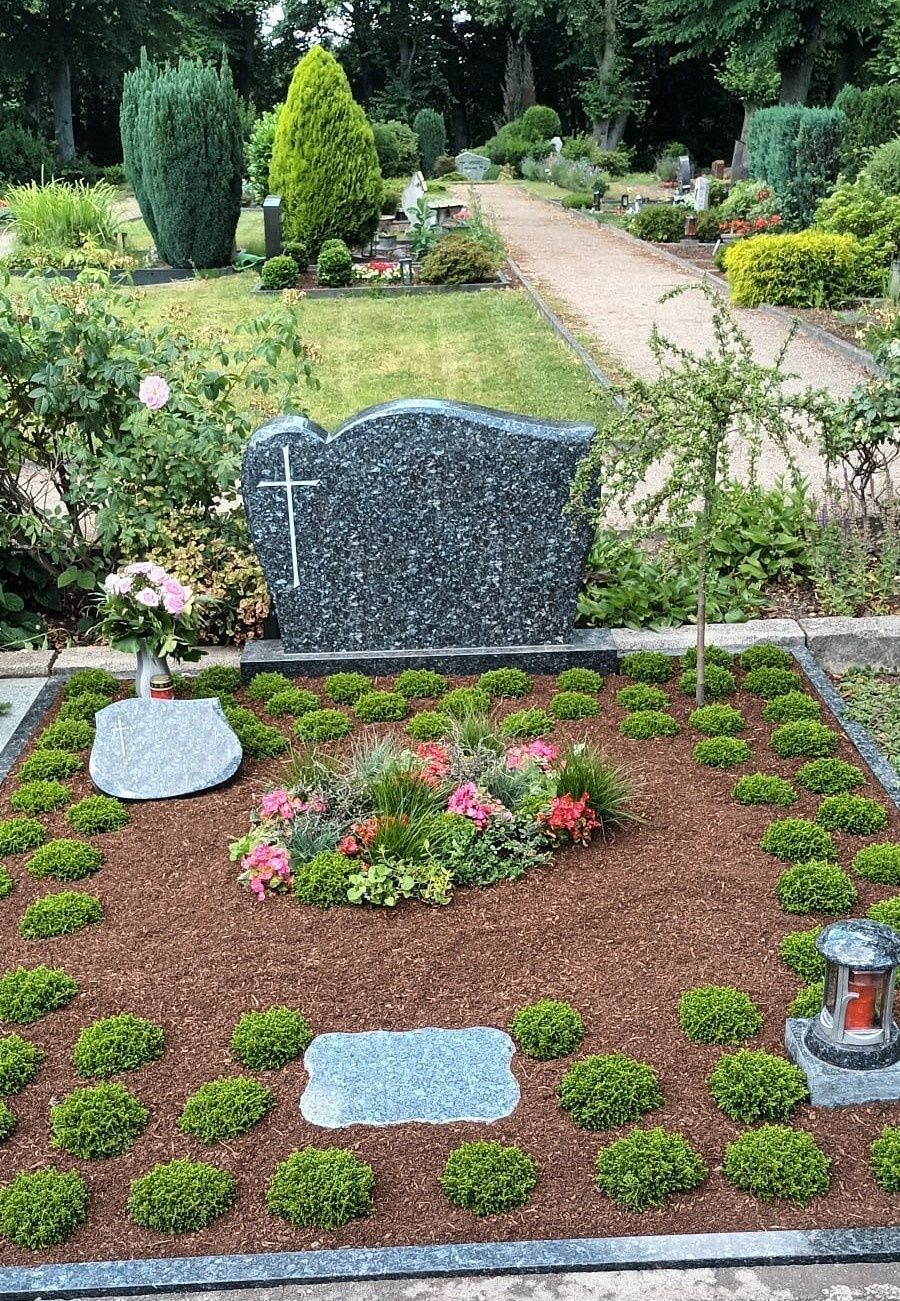 Grab mit dunklem Steingrabstein und Kreuz, umgeben von Blumen und Büschen auf einem Friedhof.