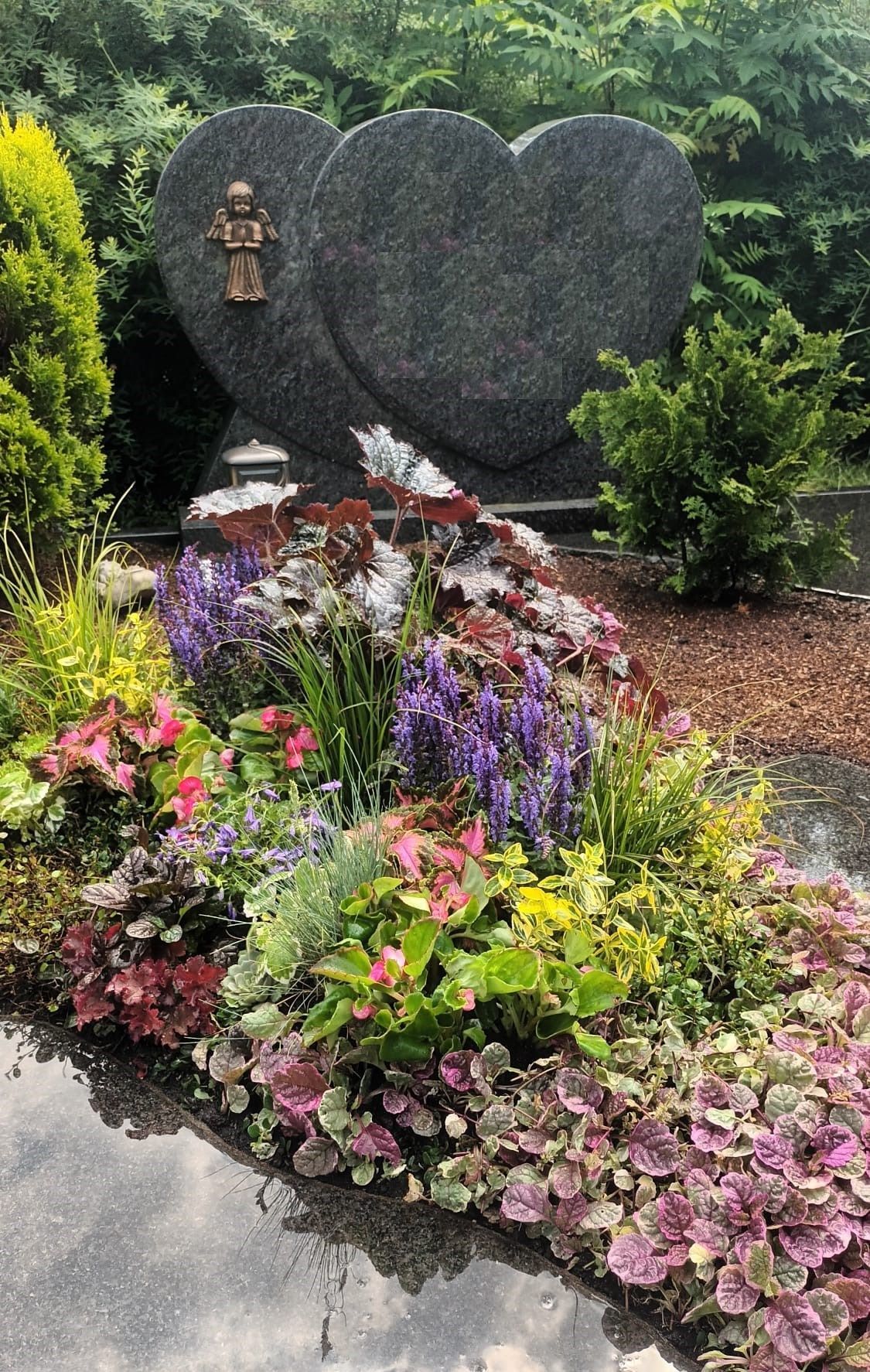 Herzförmige Grabsteine aus Granit mit einem bronzenen Kreuz, umgeben von bunten Blumen und Grünpflanzen.