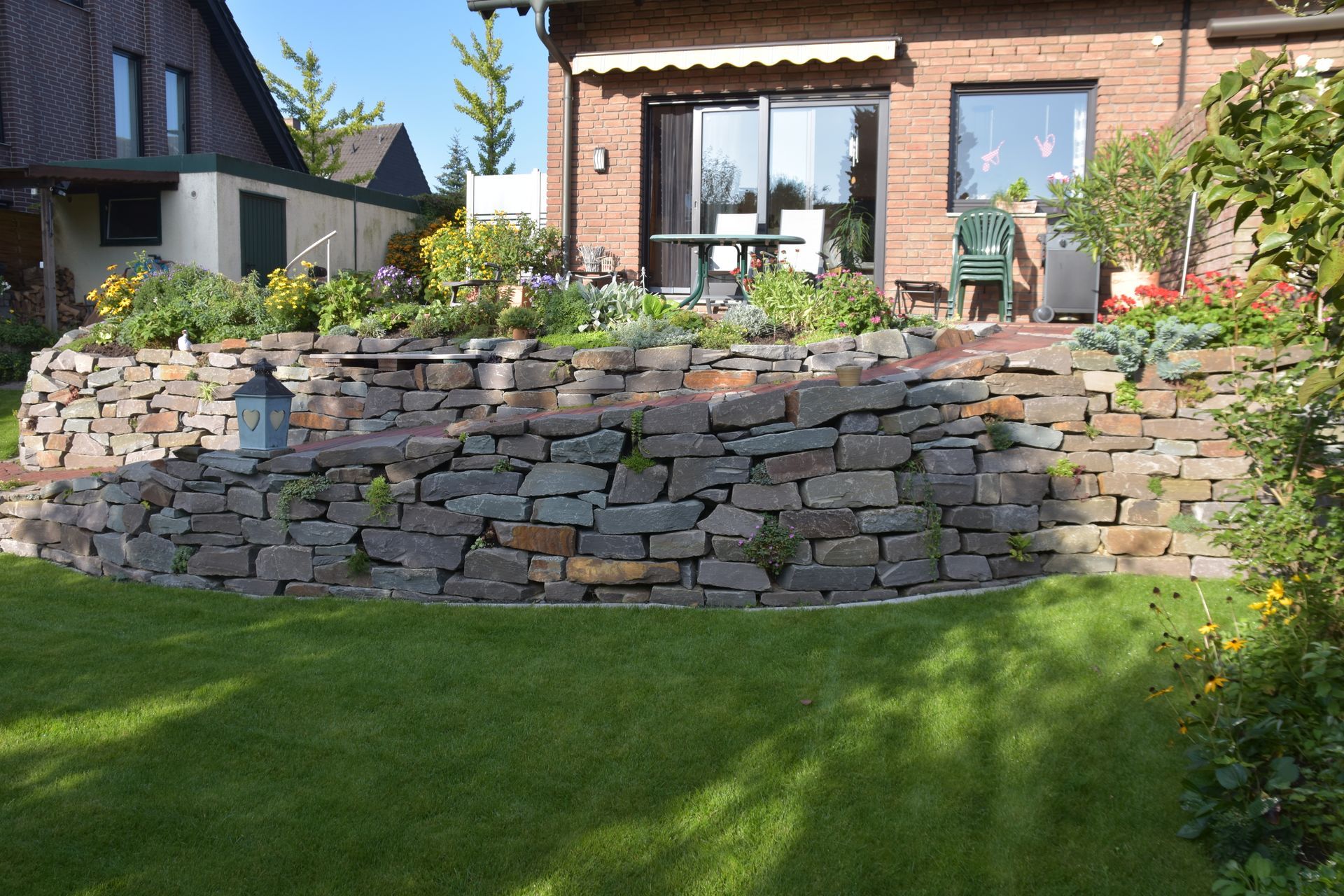 Steinstützmauer in einem Garten, die den Rasen von einem erhöhten Blumenbeet in der Nähe eines Backsteinhauses trennt.