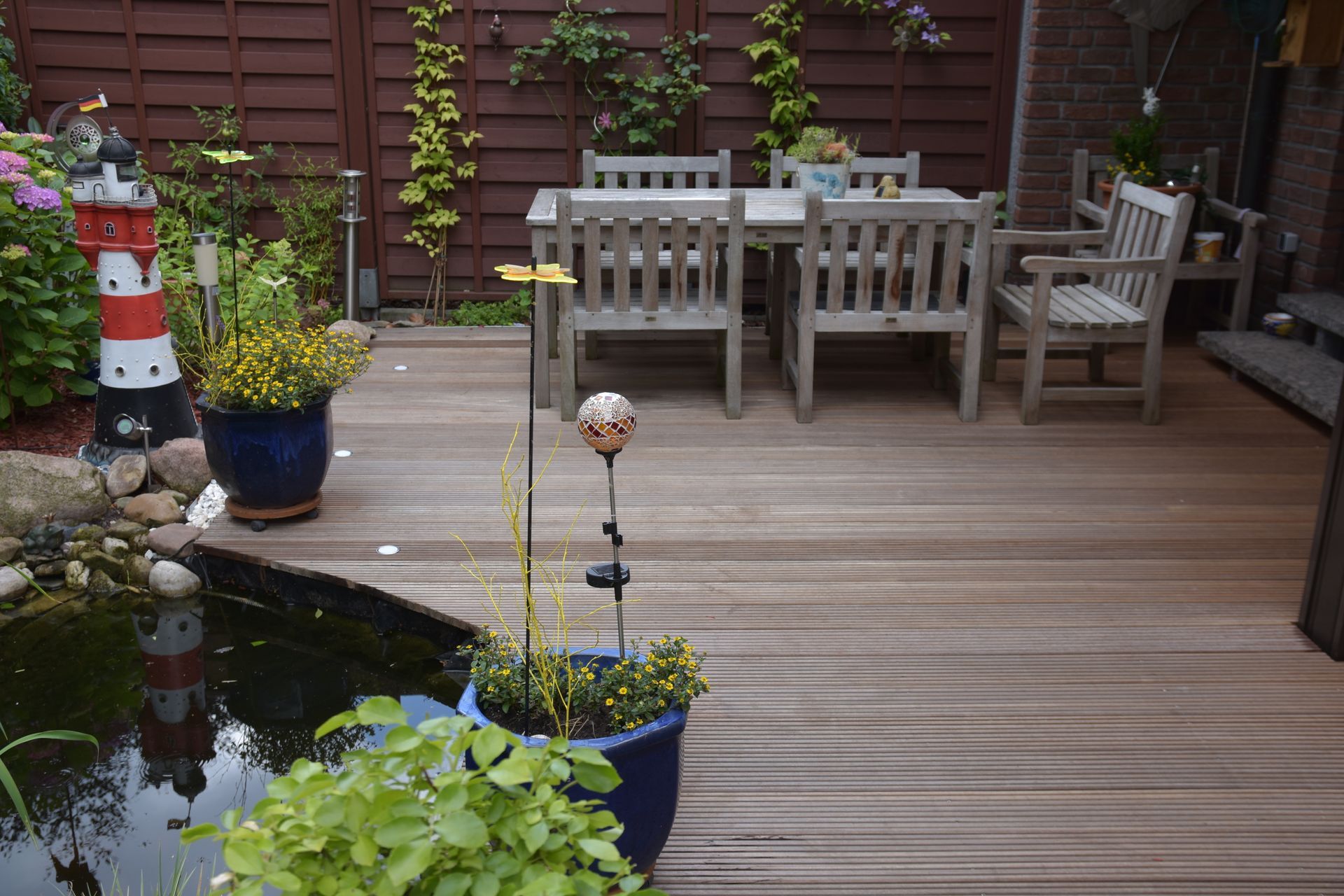 Holzterrasse mit Esstisch und Stühlen, Teich und Topfpflanzen.