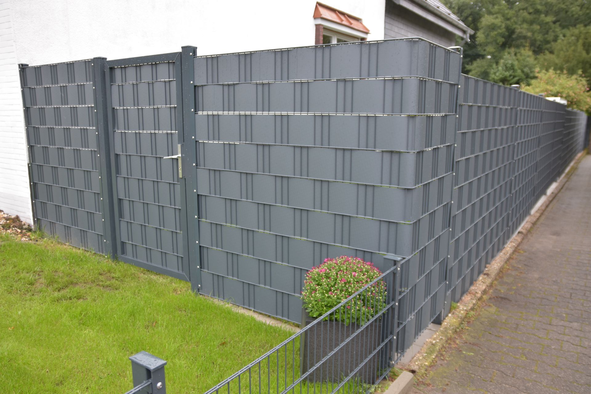 Grauer Zaun im Flechtstil mit Tor, der einen Rasenhof und eine kleine Pflanze umschließt.