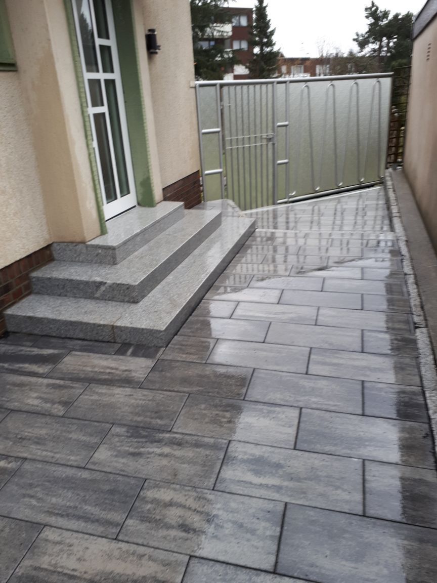 Graue Steinstufen führen zu einer Tür. Gepflasterter Gehweg neben einem Metalltor und einer Gebäudewand.