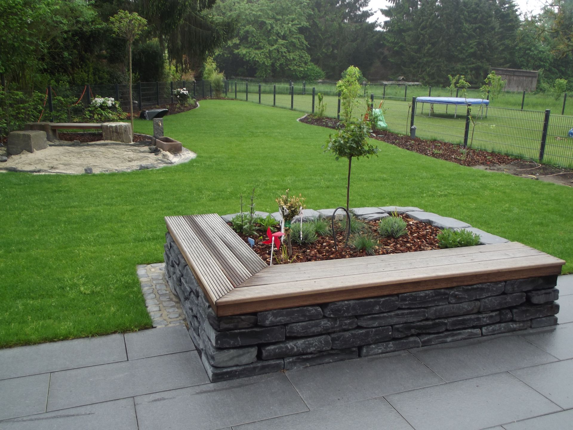 Erhöhtes Steingartenbeet mit Pflanzen, umgeben von einer Holzbank auf einem grünen Rasen.