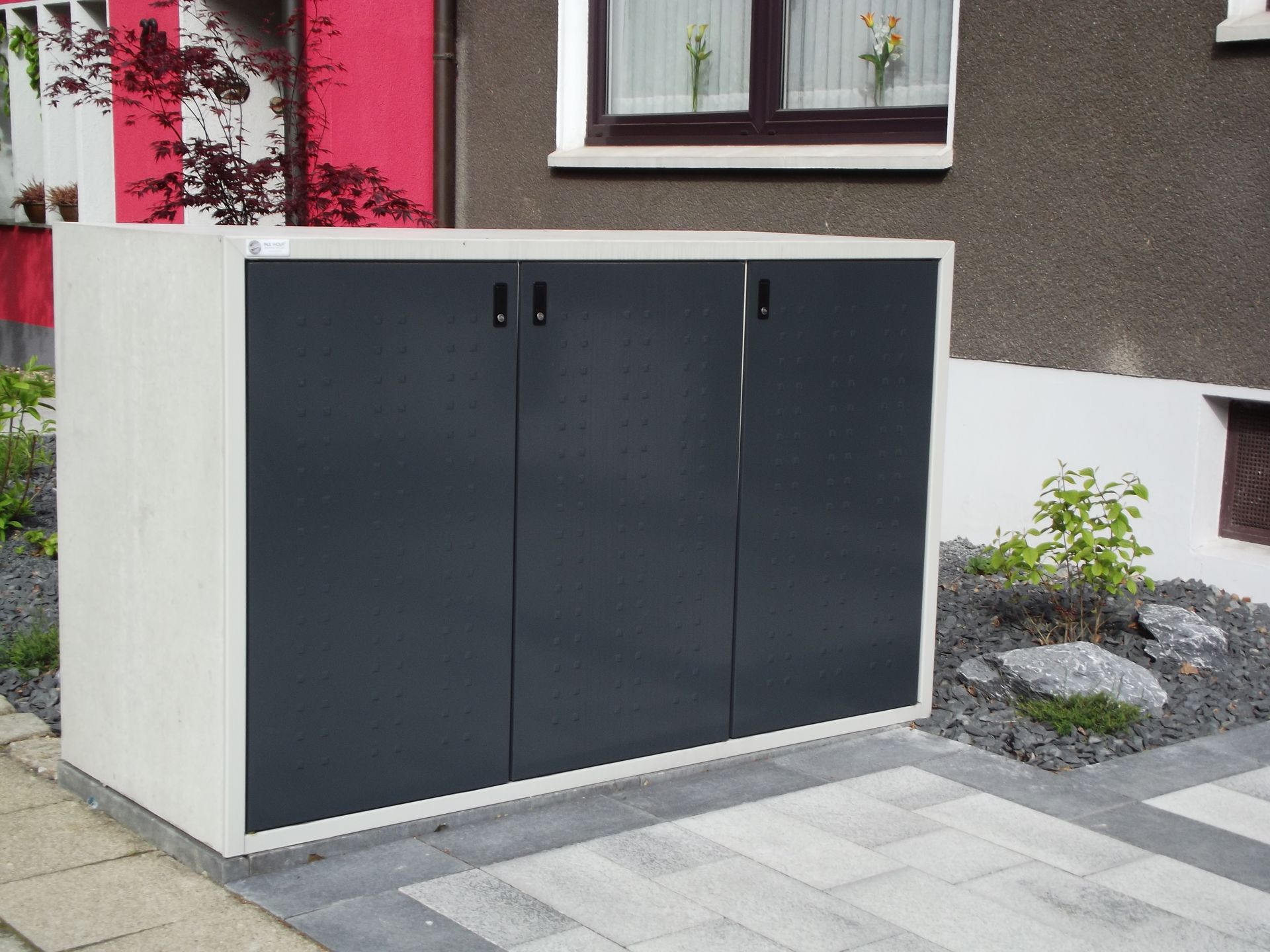 Außenlagerschrank mit drei dunkelgrauen Türen und einem hellgrauen Rahmen auf einer gepflasterten Fläche.