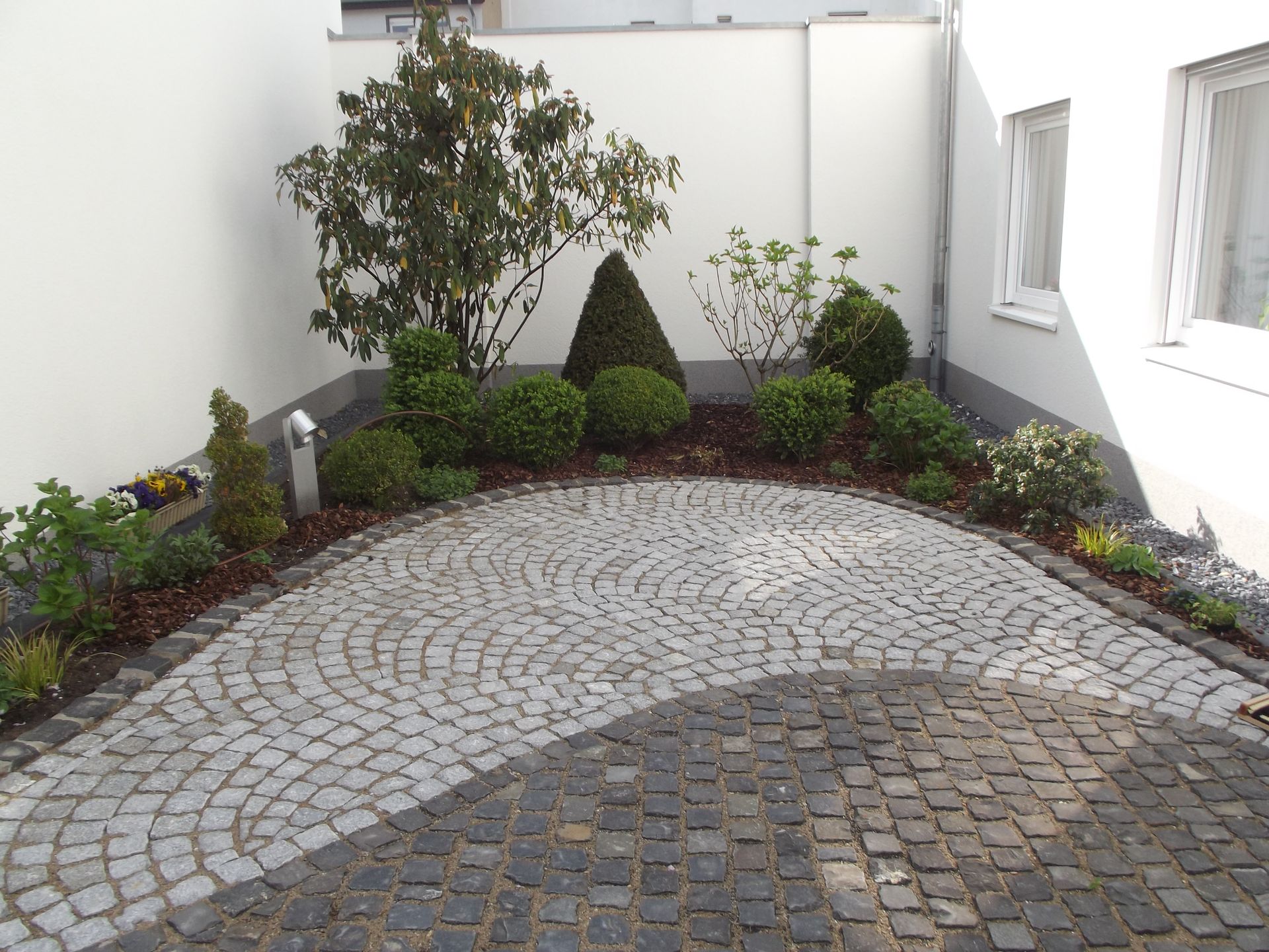 Kopfsteinpflasterterrasse mit Gartenbeet, Büschen und einem kleinen Baum vor weißen Wänden.