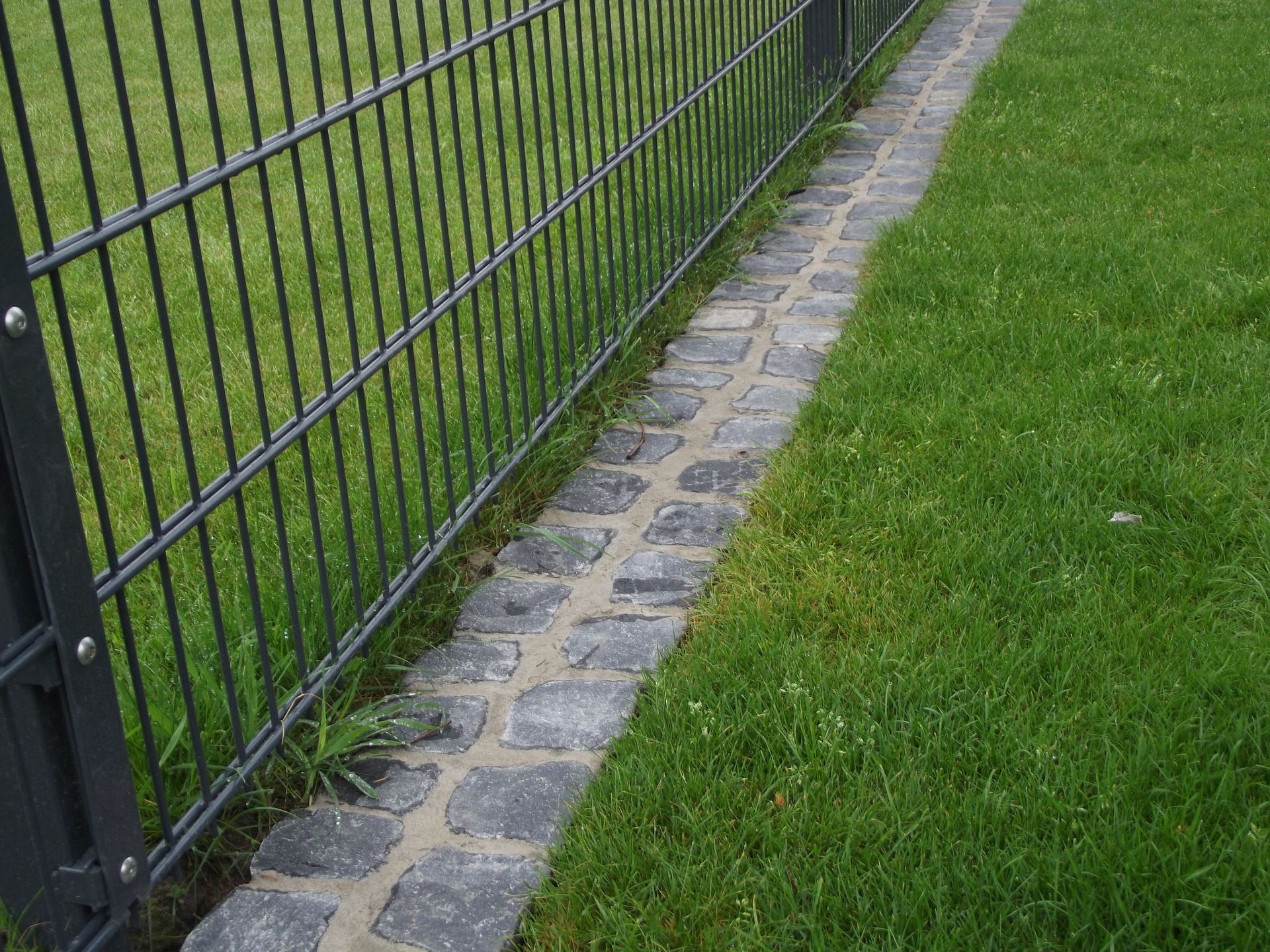 Ein schwarzer Metallzaun grenzt an einen Ziegelweg und einen grünen Rasen.
