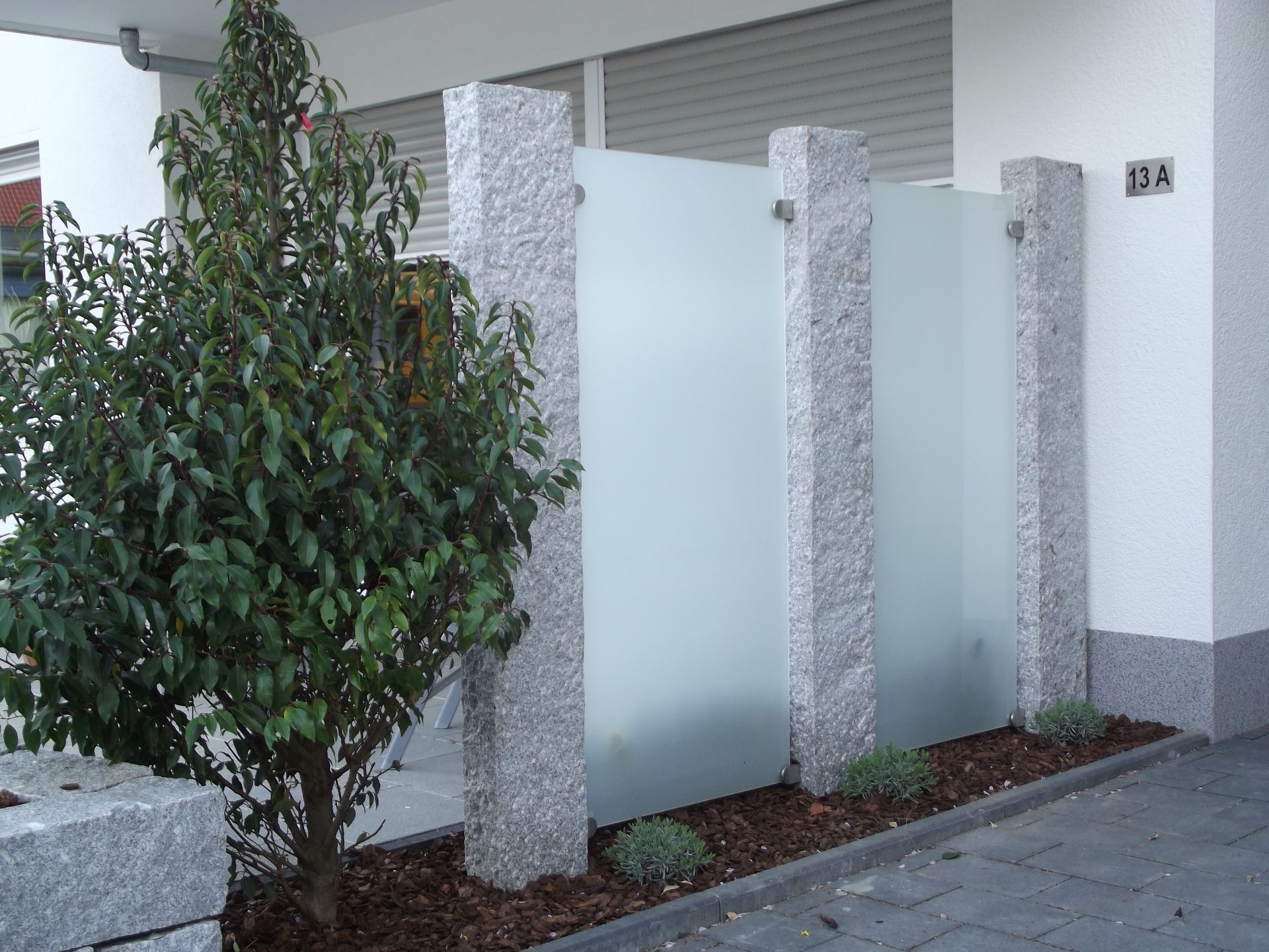 Granitpfosten stützen Milchglasplatten; ein kleiner Baum und Landschaftsgestaltung vor einem Gebäude.