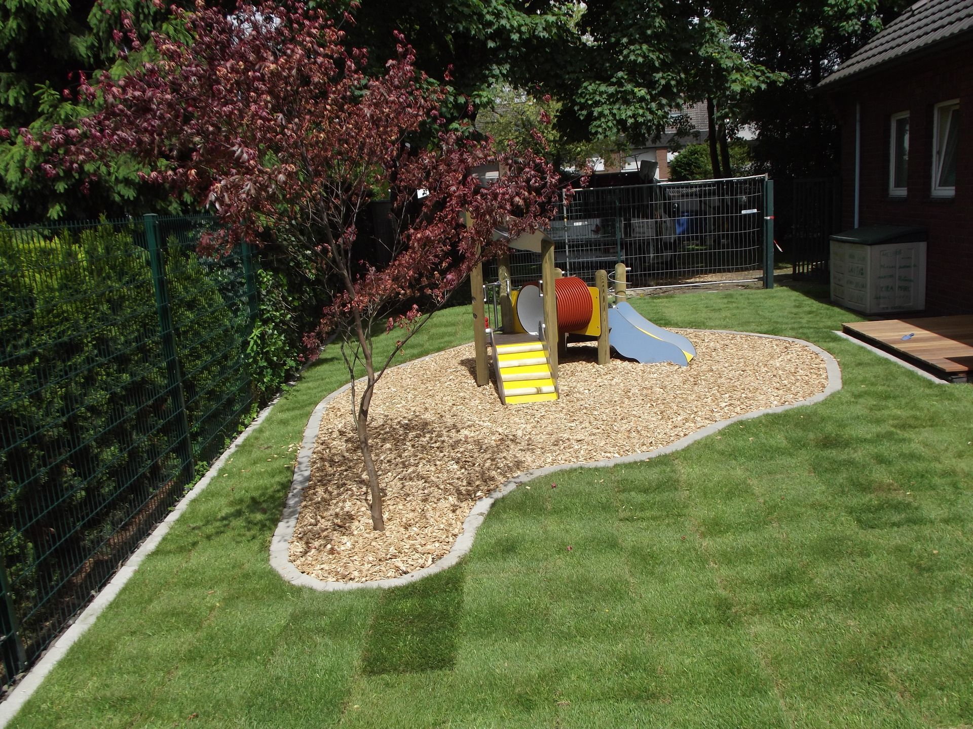 Spielplatzbereich mit hellbraunem Mulch und einem rotblättrigen Baum, umgeben von grünem Rasen und einem grünen Zaun.