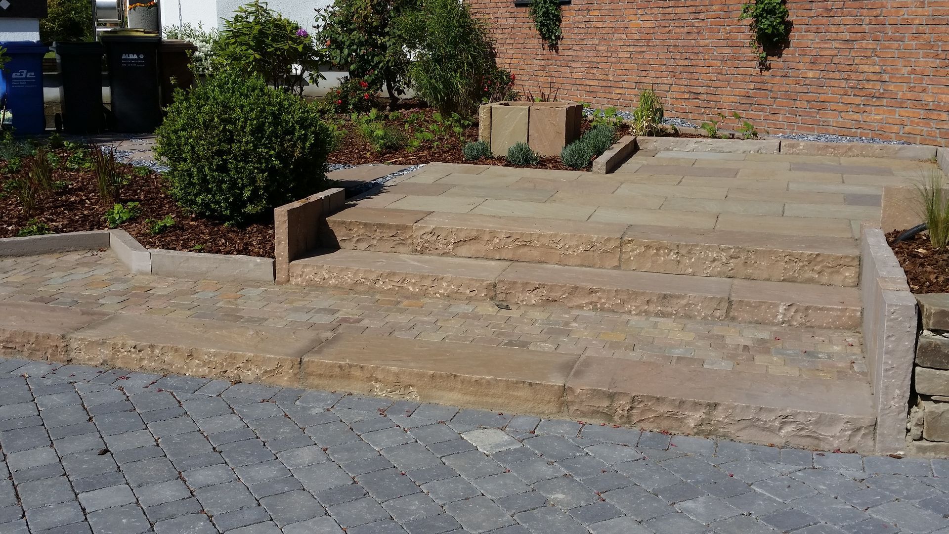 Steinstufen führen zu einem Hochbeet vor einer Ziegelmauer und grauen Pflastersteinen.