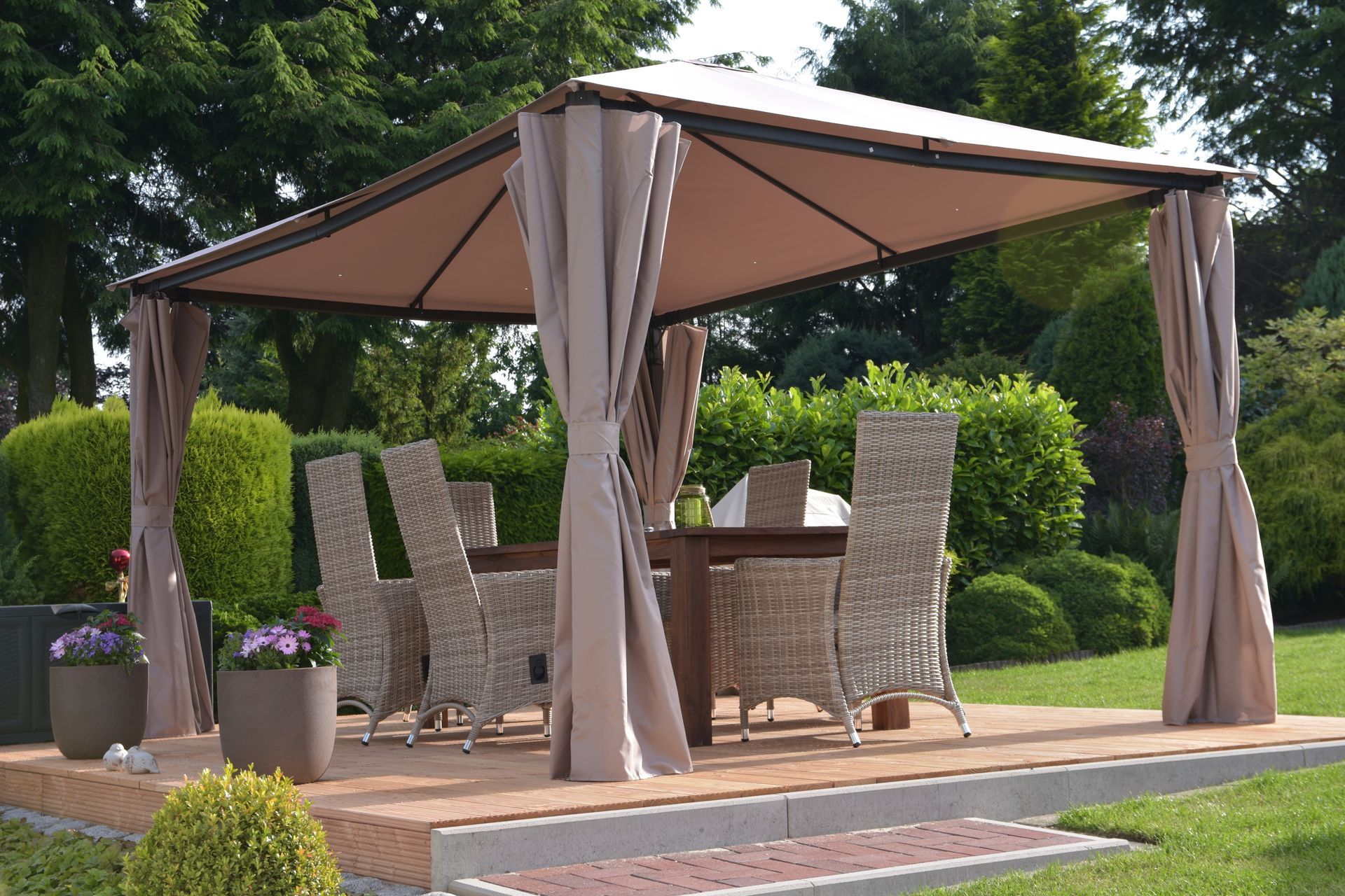 Pavillon mit Esstisch und Stühlen auf einer Terrasse. Beige Vorhänge, grünes Laub.