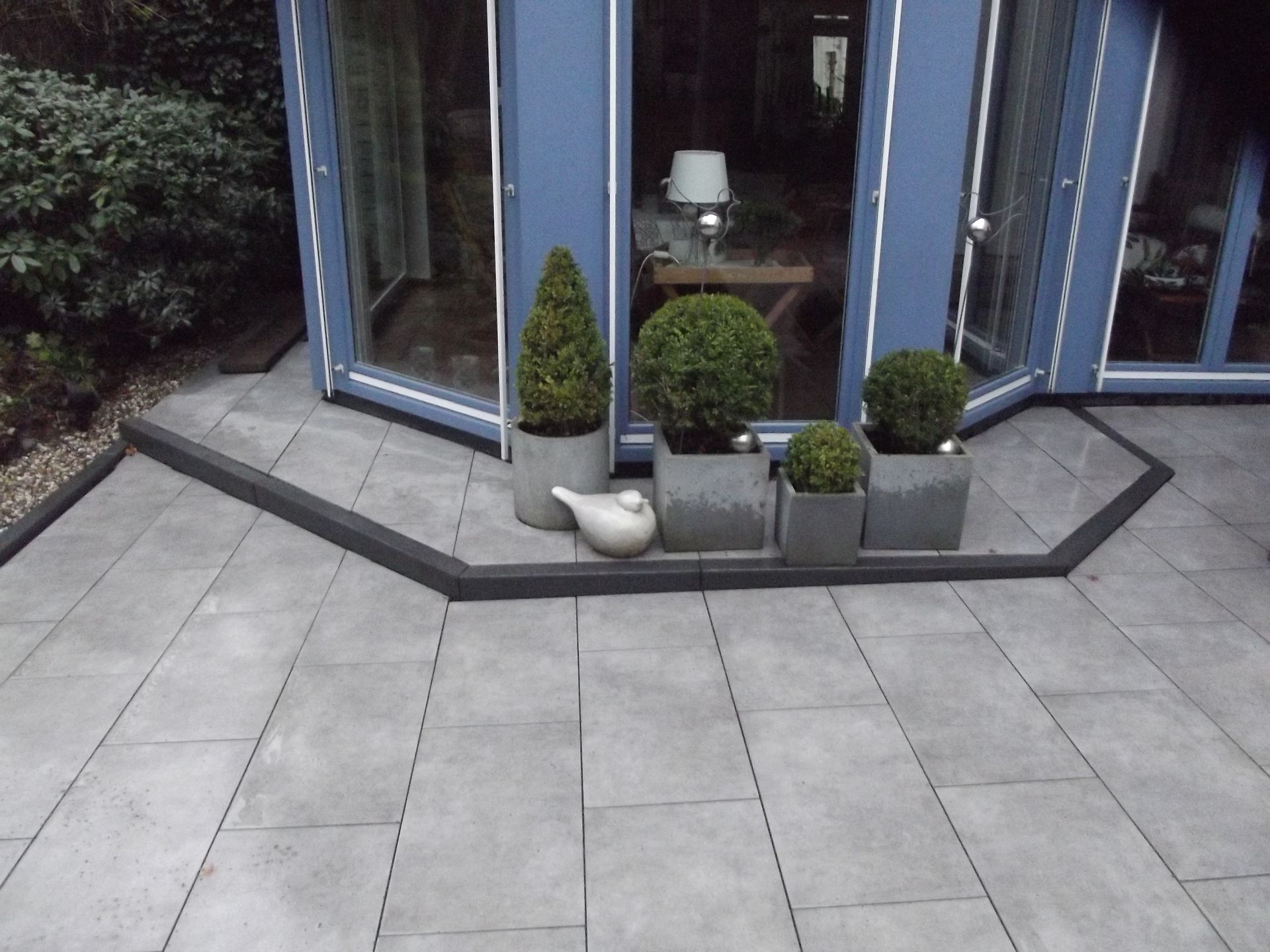 Grau gepflasterte Terrasse mit Topfpflanzen und einer Glastür mit blauem Rahmen im Hintergrund.