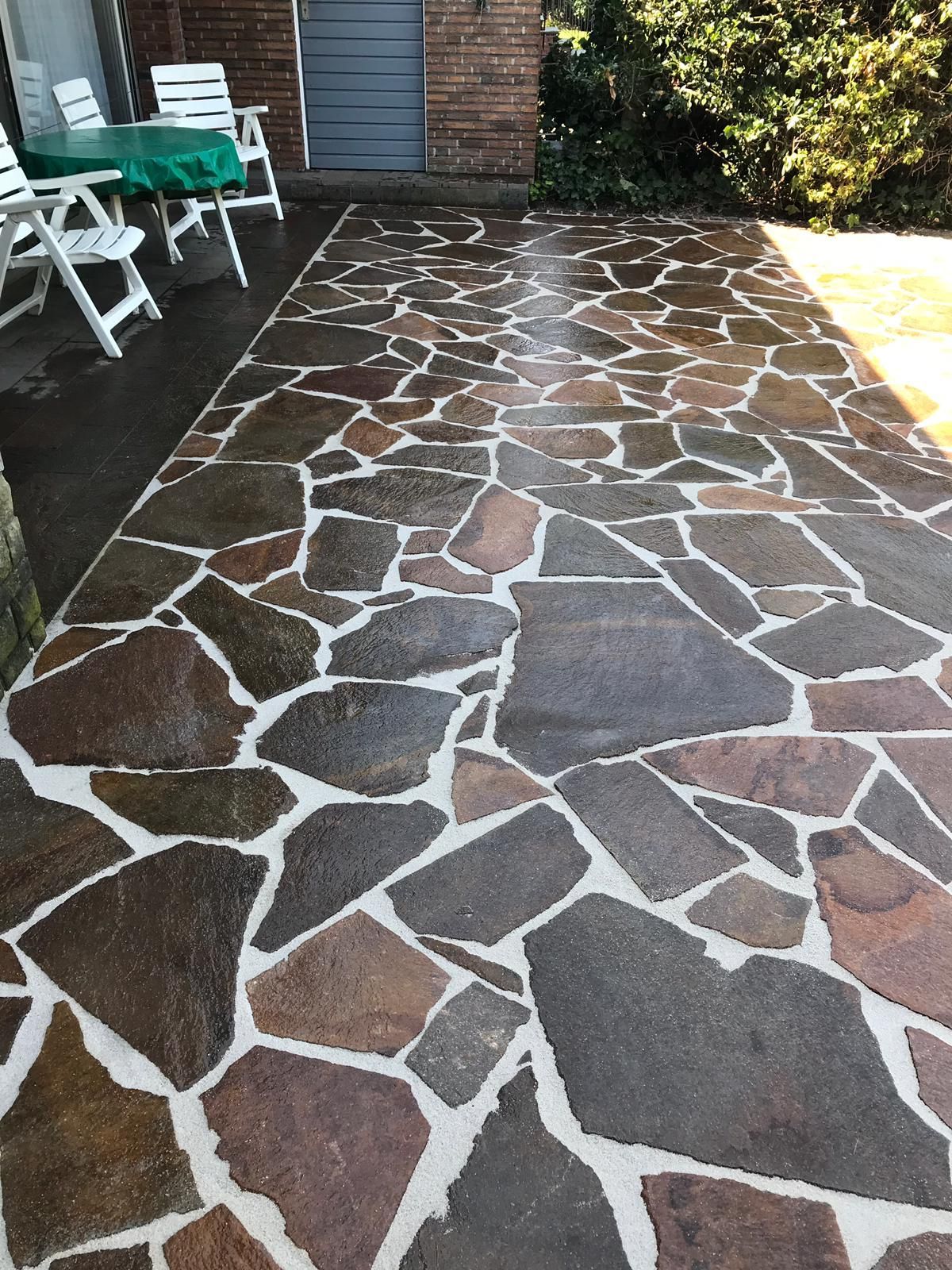 Steinplattenterrasse mit unregelmäßigen dunkel- und hellbraunen Steinen, weißem Mörtel und Gartenmöbeln.