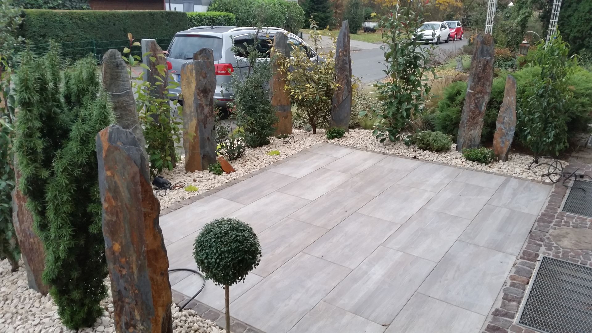 Steingarten mit großen Schieferfelsen, grauer Terrasse, Sträuchern und einem kleinen, runden Baum.