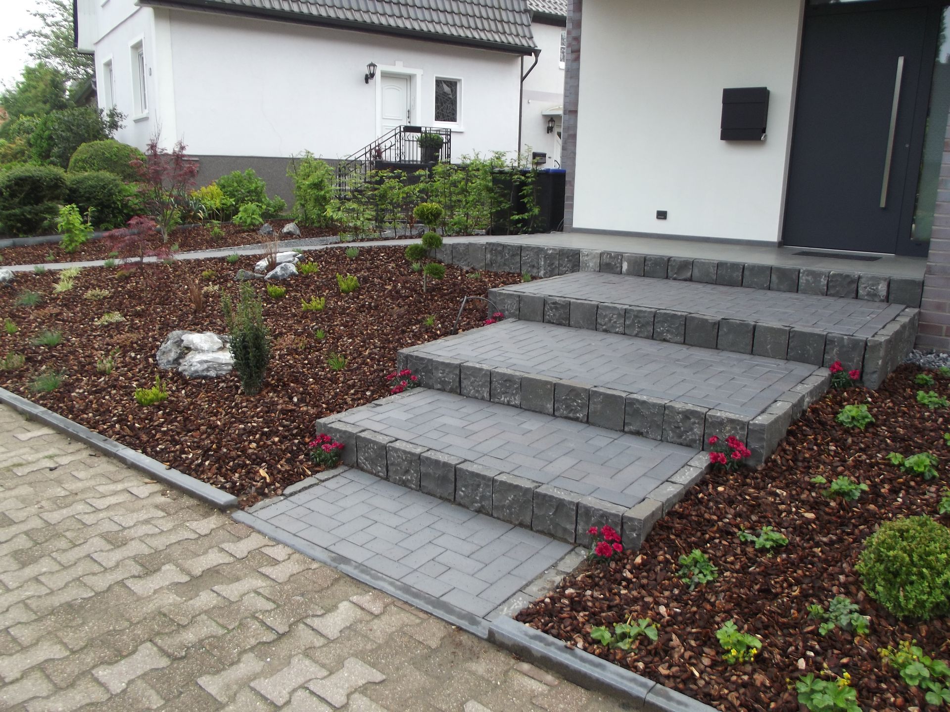Graue Ziegelstufen führen zu einem modernen Hauseingang, der von einem Garten mit Holzschnitzeln und Pflanzen begrenzt wird.