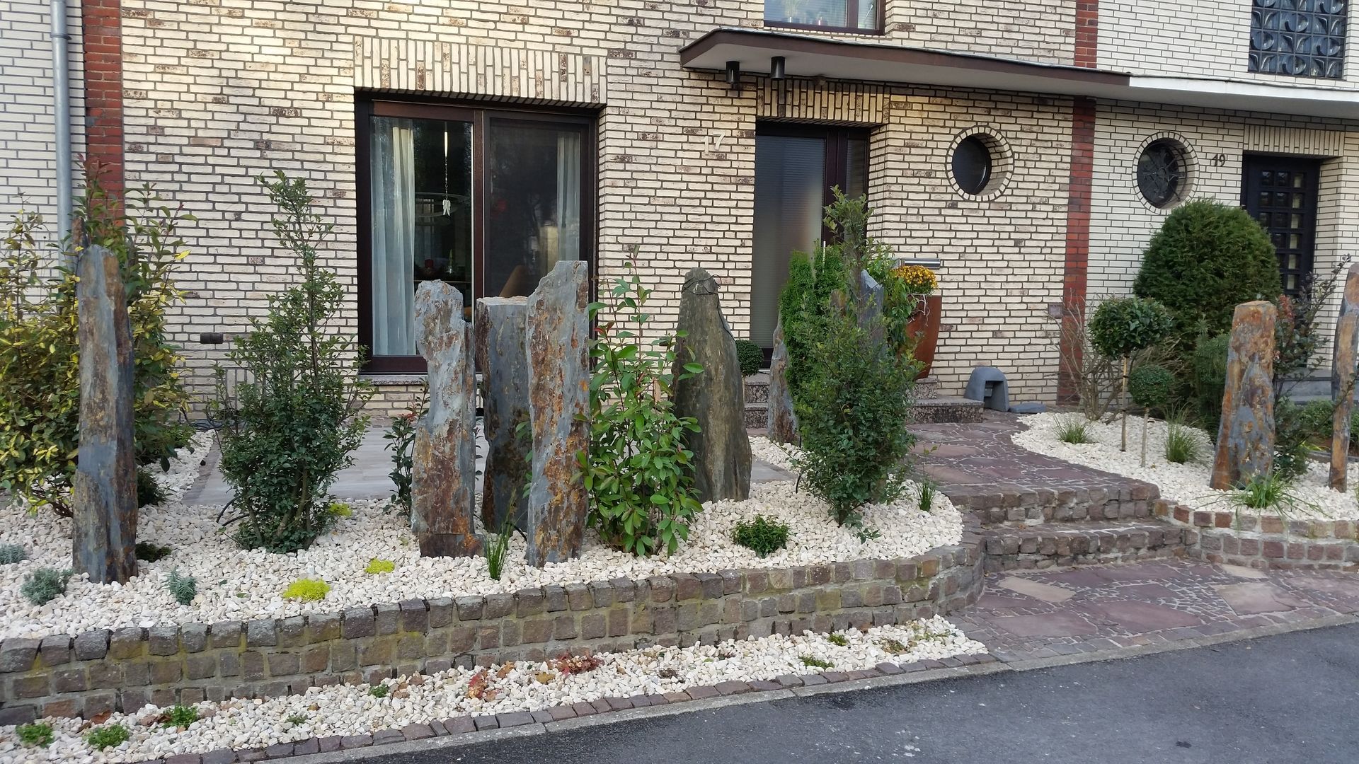 Landschaftsgestaltung des Vorgartens mit stehenden Steinskulpturen, Sträuchern und weißen Kieselsteinen.